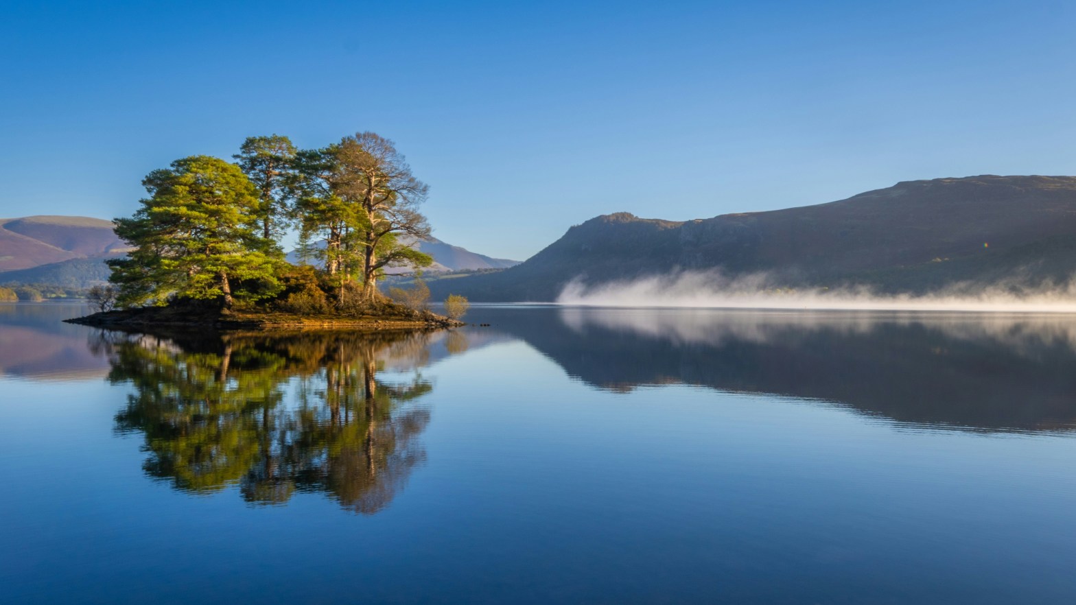 A lake with a small island and trees in the middle, reflecting in the water toward the viewer. In the background are hills and mountains.